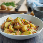 Foto von Kartoffelsalat mit Brühe und Speck auf einem Teller mit Gabel und Frühkartoffeln daneben