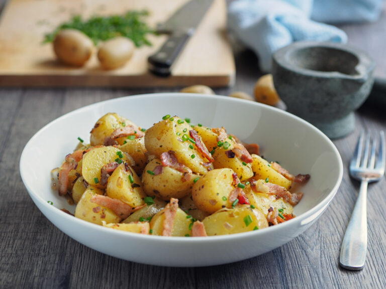 Foto von Kartoffelsalat mit Brühe und Speck auf einem Teller mit Gabel und Frühkartoffeln daneben